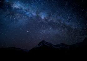 A starry night sky above dark mountain silhouettes, with one shooting star visible against the clear sky