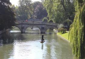 A person punting along a river toward a stone footbridge, with trees and greenery surrounding the water on a sunny day