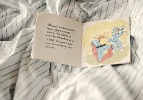 An open vintage children’s book lying on striped bed sheets, showing a scene from a British story with a teddy bear and child being launched from a toy box