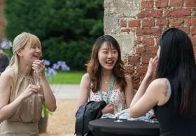 Three women are talking and laughing together outside against a red brick wall.