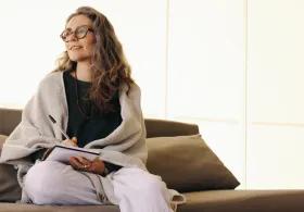A woman holding a pen poised over a notepad, sitting cross-legged on a sofa in a softly lit room