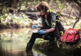 A woman testing lake water with a colour sample board, sitting on the grass with her feet in the lake, a red bag beside her and trees in the background
