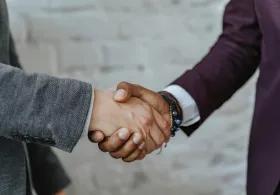 Two people in suits shaking hands, with only their hands and sleeves visible, against a blurred stone floor background