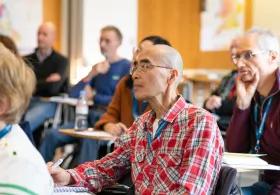 Man sitting at small table in lecture hall, other people sitting around him, paying attention to what's happening in front of them. 
