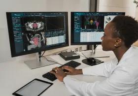 A researcher in a white coat analysing brain and body scans on dual monitors in a clinical setting
