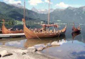 Two Viking boats near the shore with several others behind them, set against a backdrop of mountains, a lake, and a cloudy sky