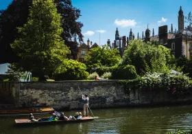 A punt full of people goes along the river cam against the backdrop of a college garden full of trees and greenery.