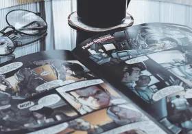 An open comic book lying on a glass table, with a black mug of coffee beside it and a pair of round glasses resting on the comic’s pages.