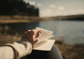 A person’s hand turning a page in a notebook while sitting by a lake, with blurred trees and sky in the background