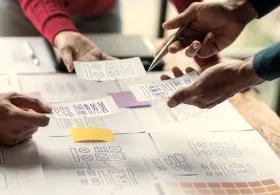 Several hands pointing to a mind map covered in post-it notes during a brainstorming session, with more notes and leaflets being held