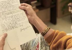 An elderly woman with glasses and a gold bracelet on her right wrist, holding and reading a handwritten letter, with the focus on the letter and her hands.
