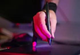A person's hand, illuminated by red light, holds a digital pen over a drawing tablet, with a black wristband visible on their wrist and a dark background. 