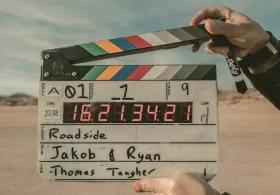 A close-up shot shows a film clapperboard being held by two hands against a blurred background of a desert landscape under an overcast sky.