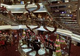 A group of people wearing Santa hats and festive attire, gathered around a grand piano and illuminated Christmas trees in the atrium of what appears to be a cruise ship, with a multi-level, mirrored staircase winding upwards in the background.