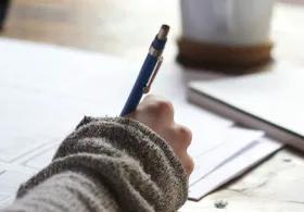 A person's hand, wearing a grey knitted sleeve, holding a blue pen and writing on a white sheet of paper on a wooden desk. A notebook and a mug are also visible in the background.