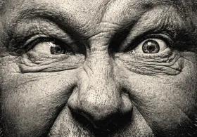 Close-up, high-contrast black and white portrait of a man with intense, wide eyes and wrinkled forehead, looking to the side with one eye and forward with the other.