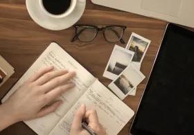 A flat lay image showing a person's hands writing in a planner or journal open to "FEBRUARY" on a wooden desk. To the left are stacked books, including one titled "YOUR SOUL" by Nikita Gill. To the right are a tablet, a pair of eyeglasses, a white coffee cup, and several small instant photo prints scattered on the desk.