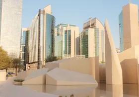 A city skyline in Abu Dhabi featuring modern skyscrapers and a park with abstract, sculptural elements and a reflective water feature, with Qasr Al Hosn visible in the background.