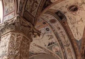 A close-up view of an elaborately decorated arched ceiling and a sculpted column inside a historic building, likely Palazzo Vecchio, showcasing intricate patterns, figures, and architectural details in warm, muted tones.