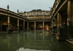 A wide shot of the Great Bath at the Roman Baths , featuring the large, green-tinged water of the main pool surrounded by ancient Roman architecture, including large columns and statues on the upper terrace, under an overcast sky."