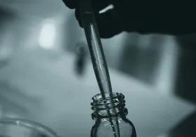  close-up, black and white image of a pipette being used to transfer liquid into a small glass bottle, likely in a laboratory or scientific setting.