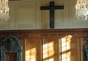 A room with wooden paneling on the lower walls and lighter-colored walls above, featuring two ornate crystal chandeliers and a large, dark crucifix mounted centrally on the upper wall. Sunlight streams through a window, casting shadows on the wooden panels.