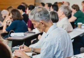 Mature male student taking notes in a lecture theatre surrounded by other students.