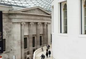 An interior shot of the British Museum's Great Court in London, featuring the vast tessellated glass roof and the circular Reading Room, with numerous visitors traversing the open space and climbing the grand staircase, visible under daylight filtering through the roof.