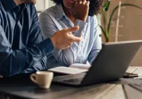 A man and a woman sitting together at an open laptop on a table, having a conversation.