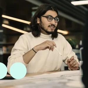 Man with shoulder length dark hair, sat at desk with pencil in hand