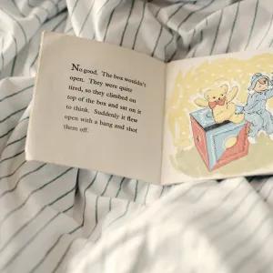 An open vintage children’s book lying on striped bed sheets, showing a scene from a British story with a teddy bear and child being launched from a toy box