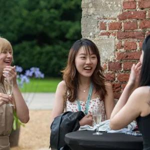 Three women are talking and laughing together outside against a red brick wall.