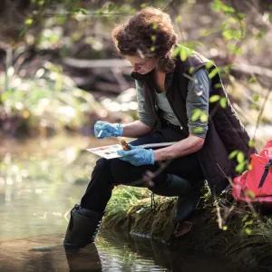 A woman testing lake water with a colour sample board, sitting on the grass with her feet in the lake, a red bag beside her and trees in the background