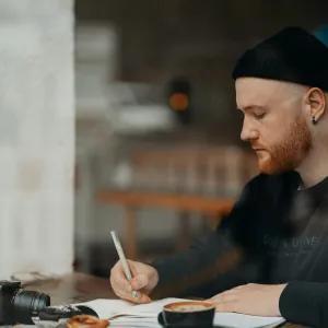 A man writing in a notebook at a table in a coffee shop, with a camera beside him and a blurred background