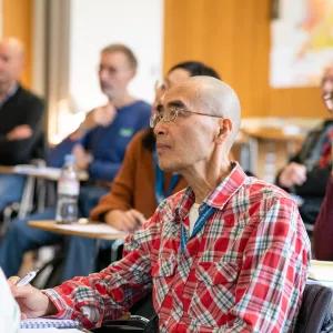 Man sitting at small table in lecture hall, other people sitting around him, paying attention to what's happening in front of them. 