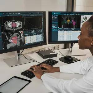A researcher in a white coat analysing brain and body scans on dual monitors in a clinical setting