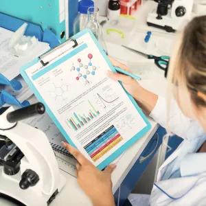 Person in lab coat looking at notes with microscope on lab bench