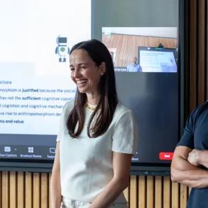 A man and a woman smiling and standing in front of a large wall-mounted display screen with a presentation on display.