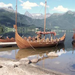 Two Viking boats near the shore with several others behind them, set against a backdrop of mountains, a lake, and a cloudy sky