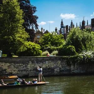 A punt full of people goes along the river cam against the backdrop of a college garden full of trees and greenery.