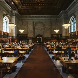People studying at desks in a long corridor of a library, surrounded by bookshelves