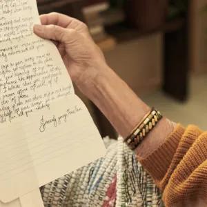 An elderly woman with glasses and a gold bracelet on her right wrist, holding and reading a handwritten letter, with the focus on the letter and her hands.