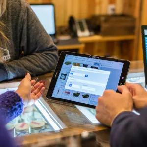 A close-up of a person holding a tablet at a shop counter, interacting with another person across the glass display. A laptop is open to a graph-based screen in the background.