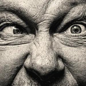 Close-up, high-contrast black and white portrait of a man with intense, wide eyes and wrinkled forehead, looking to the side with one eye and forward with the other.