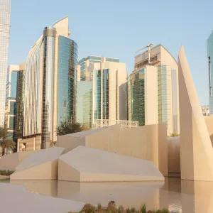 A city skyline in Abu Dhabi featuring modern skyscrapers and a park with abstract, sculptural elements and a reflective water feature, with Qasr Al Hosn visible in the background.