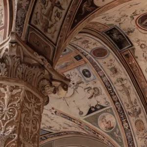 A close-up view of an elaborately decorated arched ceiling and a sculpted column inside a historic building, likely Palazzo Vecchio, showcasing intricate patterns, figures, and architectural details in warm, muted tones.