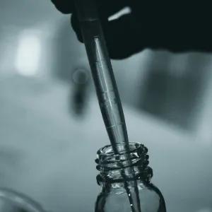  close-up, black and white image of a pipette being used to transfer liquid into a small glass bottle, likely in a laboratory or scientific setting.