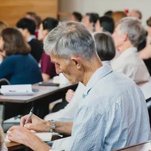 Mature male student taking notes in a lecture theatre surrounded by other students.