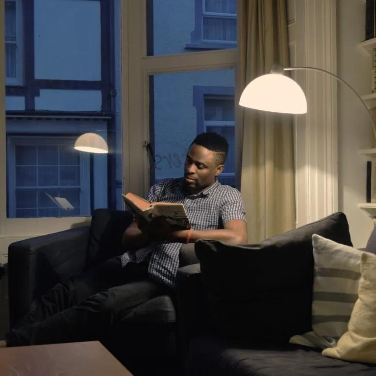 Man reading on a sofa in a dimly lit apartment, illuminated by a single overhead light