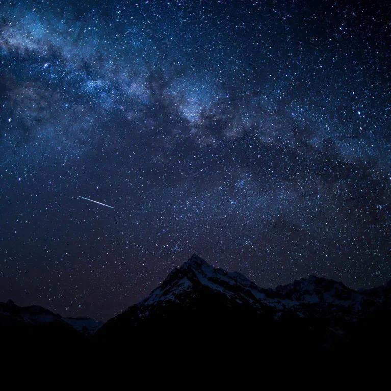 A starry night sky above dark mountain silhouettes, with one shooting star visible against the clear sky